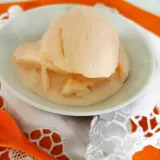 Delizioso gelato al melone solo frutta, servito in una ciotola bianca su un tovagliolo di pizzo bianco e un runner arancione. Un cucchiaio arancione è posizionato a sinistra.