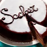 Una lucida torta Sacher, con una fetta tagliata, mostra la sua ricca copertura al cioccolato e la scritta "Sacher" decorata.