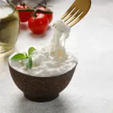 Una ciotola di stracciatella fatta in casa, con pomodorini e olio d'oliva sullo sfondo. Una forchetta solleva fili di formaggio fresco.