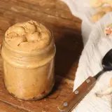 Barattolo di burro di arachidi fatto in casa, pronto per la tua prossima ricetta. Arachidi e cucchiaio su sfondo rustico.