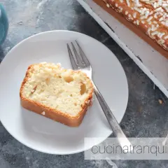 Una fetta di plumcake yogurt greco, soffice e cosparso di zuccherini, attende su un piatto bianco, accanto a una tazza di tè.