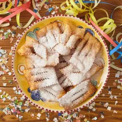Chiacchiere di Carnevale Benedetta Rossi, cosparse di zucchero a velo, su un piatto decorato, circondate da coriandoli e stelle filanti.