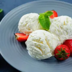 Tre palline di cremoso gelato alla ricotta, guarnite con fragole fresche e una fogliolina di menta, su un piatto grigio.