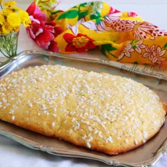 Una ciambella romagnola, cosparsa di zuccherini, su un vassoio d'argento, con fiori gialli e un foulard colorato sullo sfondo.
