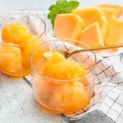 Sorbetto melone fresco e dissetante, servito in coppette di vetro con cucchiaini dorati. Fette di melone e menta sullo sfondo.