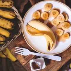 Un cestino di banane mature accanto a una banana sbucciata e affettata su un piatto, con sciroppo. Un'idea per sostituire lo zucchero con la banana.