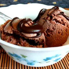 Due palline di gelato al cioccolato vegano, guarnite con sciroppo al cioccolato, in una ciotola bianca con fiori azzurri.