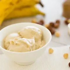 Una ciotola di cremoso gelato alla banana, con banane mature sullo sfondo e nocciole sparse.