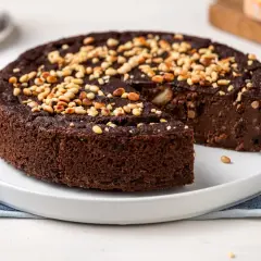 Torta paesana al cioccolato, ricca e umida, guarnita con pinoli tostati. Una fetta è stata già servita, invitando a gustare questo dolce rustico.