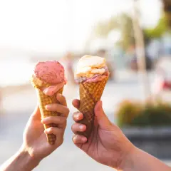 Due mani sollevano due coni gelato, uno rosa e uno con sfumature crema e rosa, celebrando la giornata del gelato.