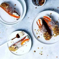Tre cannoli siciliani, guarniti con crema, arancia candita e pistacchi tritati, su piatti bianchi, spolverati di zucchero a velo.