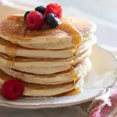 Pila di soffici pancake senza glutine, guarniti con frutti di bosco e sciroppo d'acero che cola.