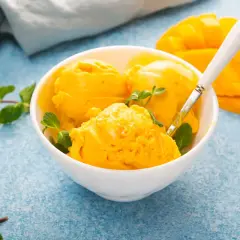 Coppa di sorbetto al mango con menta fresca e una fetta di mango sullo sfondo.