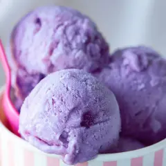 Tre palline di gelato viola in una coppetta a righe rosa e bianche, con un cucchiaino rosa. Un dolce piacere che non causa stitichezza.