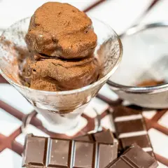 Coppetta di gelato al cioccolato spolverata di cacao, con tavolette di cioccolato e colino sullo sfondo.