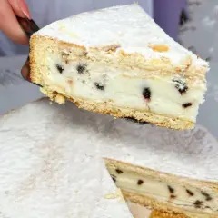 Una fetta di cassata al forno, ricca di crema e gocce di cioccolato, viene sollevata da una torta intera cosparsa di zucchero a velo.