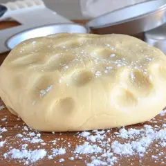 Impasto di pasta frolla pronto per essere lavorato, cosparso di zucchero a velo su un tagliere di legno. Attrezzi da cucina sullo sfondo.