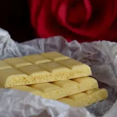 Barrette di cioccolato bianco, con la loro texture ariosa, avvolte in carta crespa, sullo sfondo di una rosa rossa. Ecco com'è fatto il cioccolato bianco.
