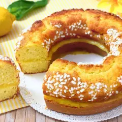 Ciambellone al limone soffice e profumato, con glassa gialla e zuccherini bianchi. Due limoni freschi e una foglia verde completano la scena.