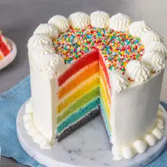 Torta arcobaleno con strati colorati, glassa bianca e zuccherini. Una fetta mostra i colori vivaci, come un vero arcobaleno.