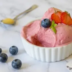 Delizioso gelato rosa, guarnito con mirtilli e fragole, servito in una ciotola bianca. Intorno, altri mirtilli e un cucchiaino con un po' di miele. Perfetto per un momento di fresco piacere, questo gelato palline di ghiaccio è un sogno.