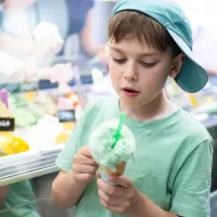 Bambino con cappellino azzurro gusta un gelato verde, pensando a chi ha inventato questa delizia.