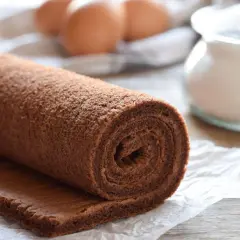 Rotolo di pasta biscotto al cacao, pronto per essere farcito. Ingredienti freschi come uova e zucchero sullo sfondo.