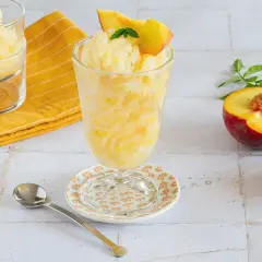 Rinfrescante granita alla pesca, guarnita con una fetta di pesca e menta. Accanto, pesche fresche e un cucchiaino.