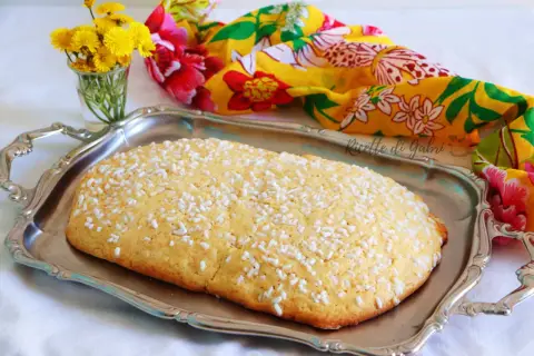Una ciambella romagnola, cosparsa di zuccherini, su un vassoio d'argento, con fiori gialli e un foulard colorato sullo sfondo.