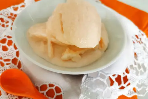 Delizioso gelato al melone solo frutta, servito in una ciotola bianca su un tovagliolo di pizzo bianco e un runner arancione. Un cucchiaio arancione è posizionato a sinistra.