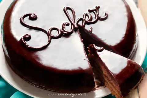 Una lucida torta Sacher, con una fetta tagliata, mostra la sua ricca copertura al cioccolato e la scritta "Sacher" decorata.