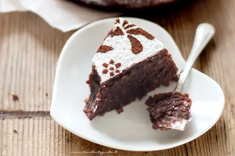 Una fetta di torta caprese ricetta originale napoletana, cosparsa di zucchero a velo e cacao, su un piatto bianco a forma di cuore.