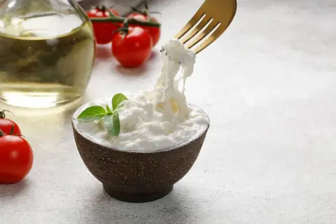 Una ciotola di stracciatella fatta in casa, con pomodorini e olio d'oliva sullo sfondo. Una forchetta solleva fili di formaggio fresco.