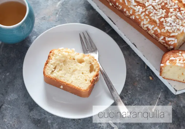 Una fetta di plumcake yogurt greco, soffice e cosparso di zuccherini, attende su un piatto bianco, accanto a una tazza di tè.