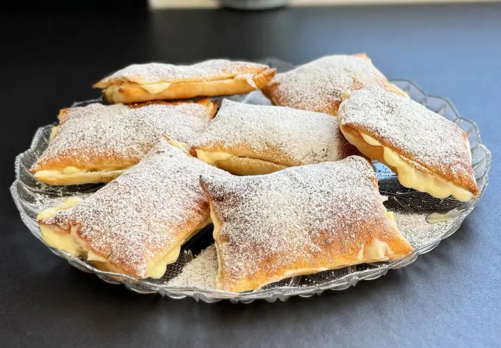 Deliziosi dolci con pasta sfoglia ripieni di crema e spolverati di zucchero a velo, serviti su un piatto di vetro.