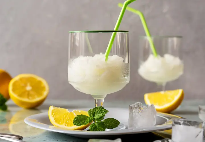 Sorbetto al limone fresco, preparato con la gelatiera, servito in bicchieri con cannucce verdi, guarnito con spicchi di limone e menta.
