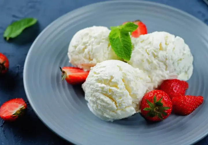 Tre palline di cremoso gelato alla ricotta, guarnite con fragole fresche e una fogliolina di menta, su un piatto grigio.