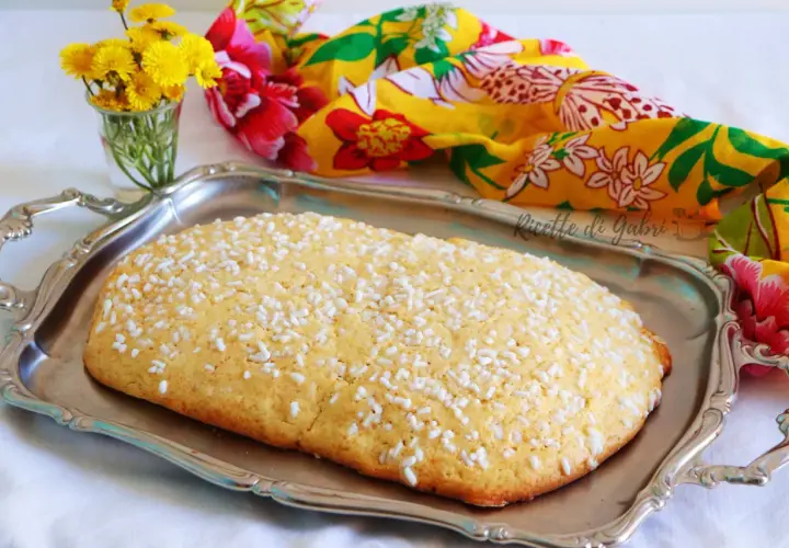 Una ciambella romagnola, cosparsa di zuccherini, su un vassoio d'argento, con fiori gialli e un foulard colorato sullo sfondo.