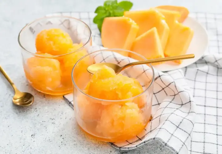 Sorbetto melone fresco e dissetante, servito in coppette di vetro con cucchiaini dorati. Fette di melone e menta sullo sfondo.