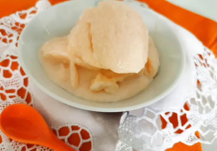 Delizioso gelato al melone solo frutta, servito in una ciotola bianca su un tovagliolo di pizzo bianco e un runner arancione. Un cucchiaio arancione è posizionato a sinistra.
