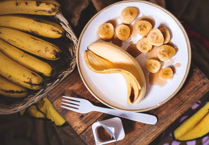 Un cestino di banane mature accanto a una banana sbucciata e affettata su un piatto, con sciroppo. Un'idea per sostituire lo zucchero con la banana.