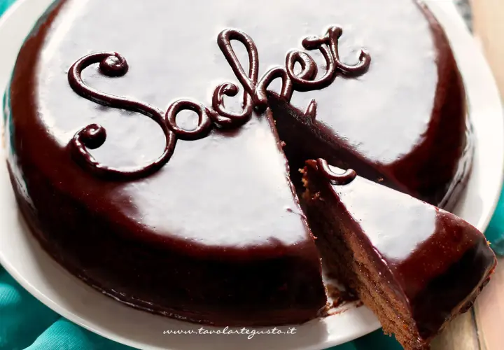 Una lucida torta Sacher, con una fetta tagliata, mostra la sua ricca copertura al cioccolato e la scritta "Sacher" decorata.