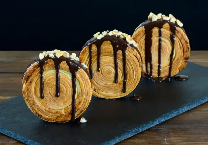 Tre deliziosi new york roll, ricoperti di cioccolato fuso e granella di nocciole, su una lavagna scura.