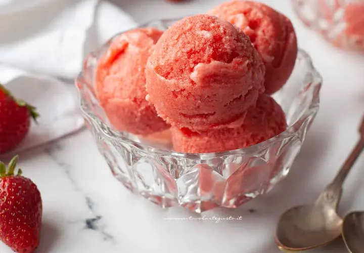 Coppa di sorbetto cremoso alle fragole, fresca e invitante, con fragole fresche e cucchiaini d'argento su sfondo marmorizzato.