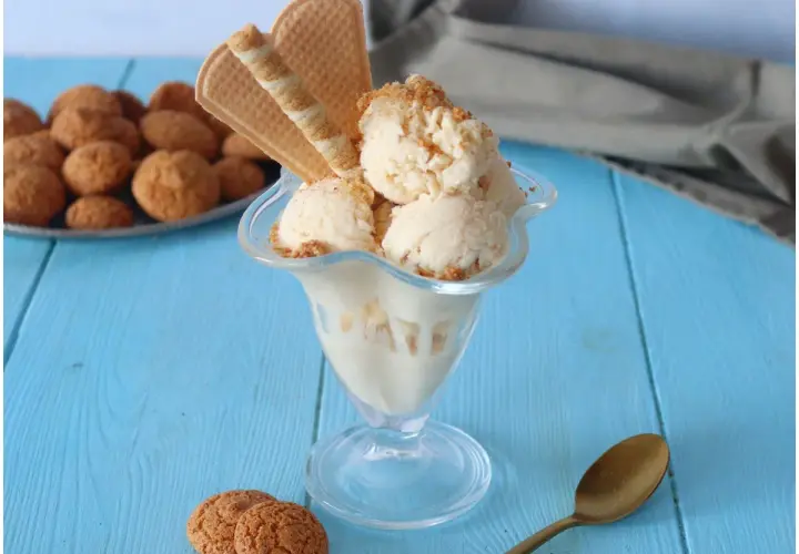 Coppa di gelato amaretto con biscotti e cialde su tavolo blu.