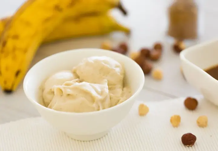 Una ciotola di cremoso gelato alla banana, con banane mature sullo sfondo e nocciole sparse.