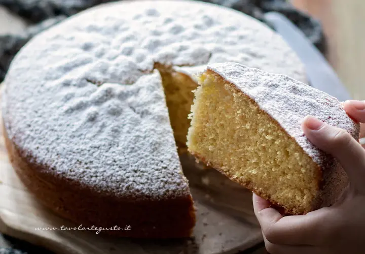 Una fetta di soffice torta margherita, cosparsa di zucchero a velo, viene sollevata da una mano.