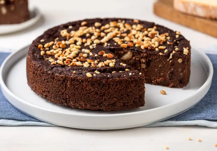 Torta paesana al cioccolato, ricca e umida, guarnita con pinoli tostati. Una fetta è stata già servita, invitando a gustare questo dolce rustico.
