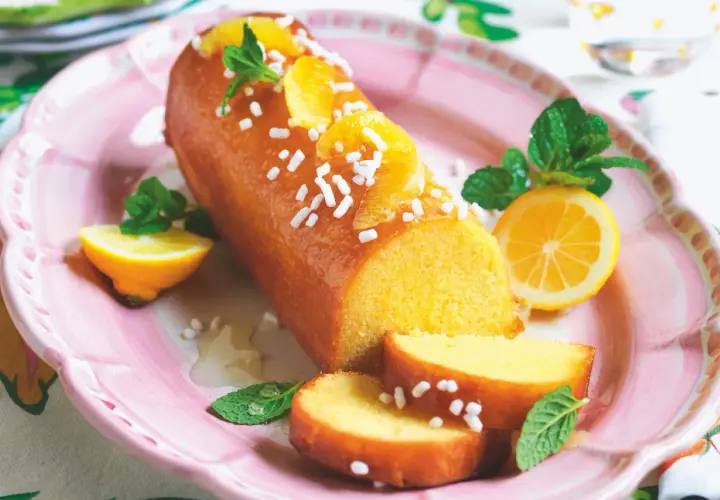 Una deliziosa torta al limone glassata, decorata con fette d'arancia, zuccherini e menta, servita su un piatto rosa con spicchi di limone.