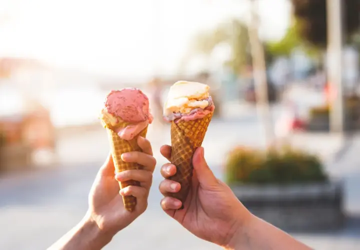 Due mani sollevano due coni gelato, uno rosa e uno con sfumature crema e rosa, celebrando la giornata del gelato.